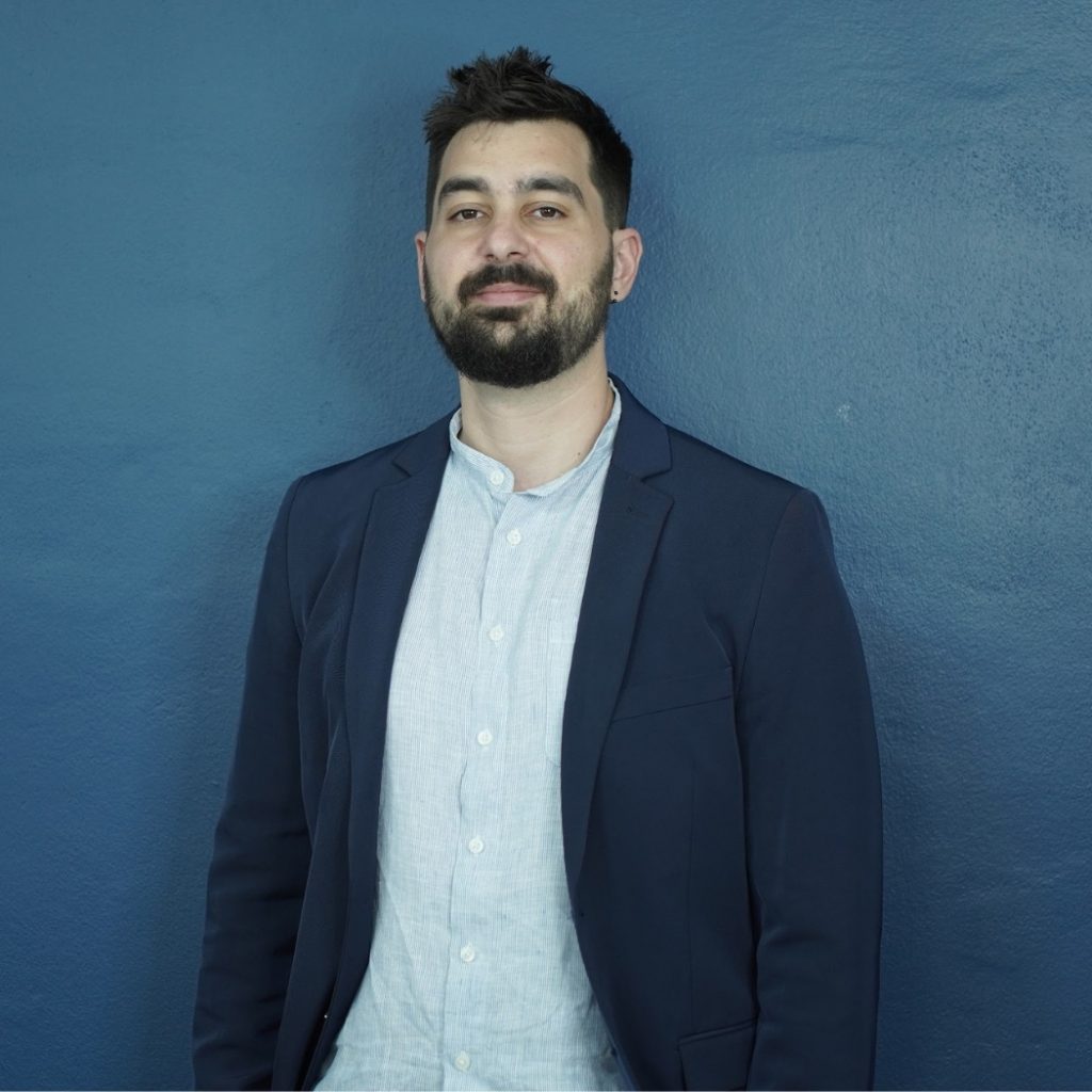 Portrait of a team member with short dark hair and a beard, wearing a light shirt and navy blazer, standing against a textured dark blue wall.