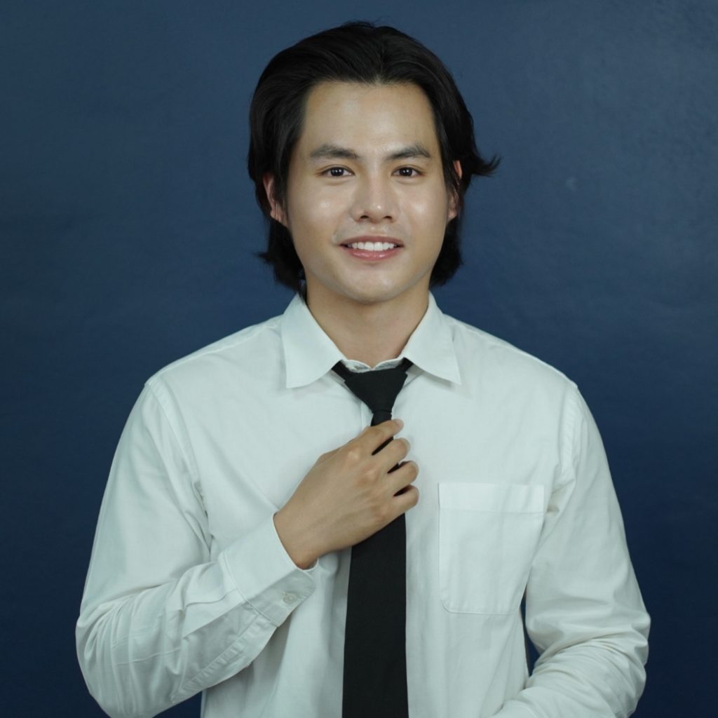 Portrait of a team member adjusting his black tie while wearing a white shirt, smiling in front of a dark blue background.