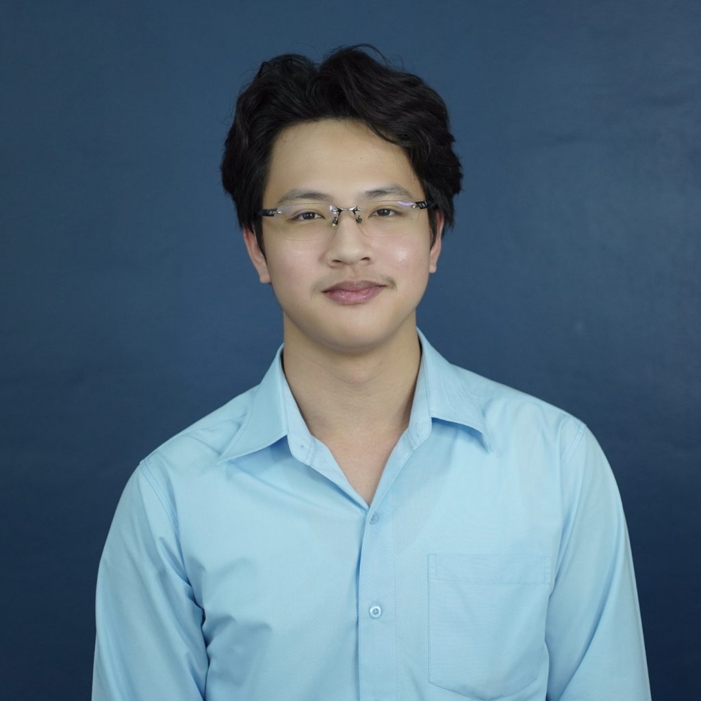 Portrait of a team member wearing glasses and a light blue shirt, standing in front of a dark blue background.